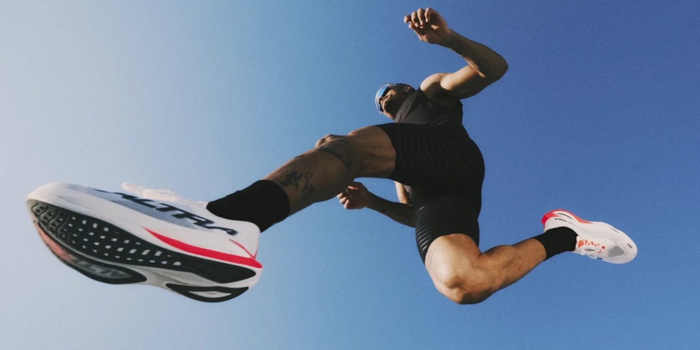 Male runner leaping skyward wearing white and red Altra Vanish Carbon 3 racing shoes, shot from below against blue sky.