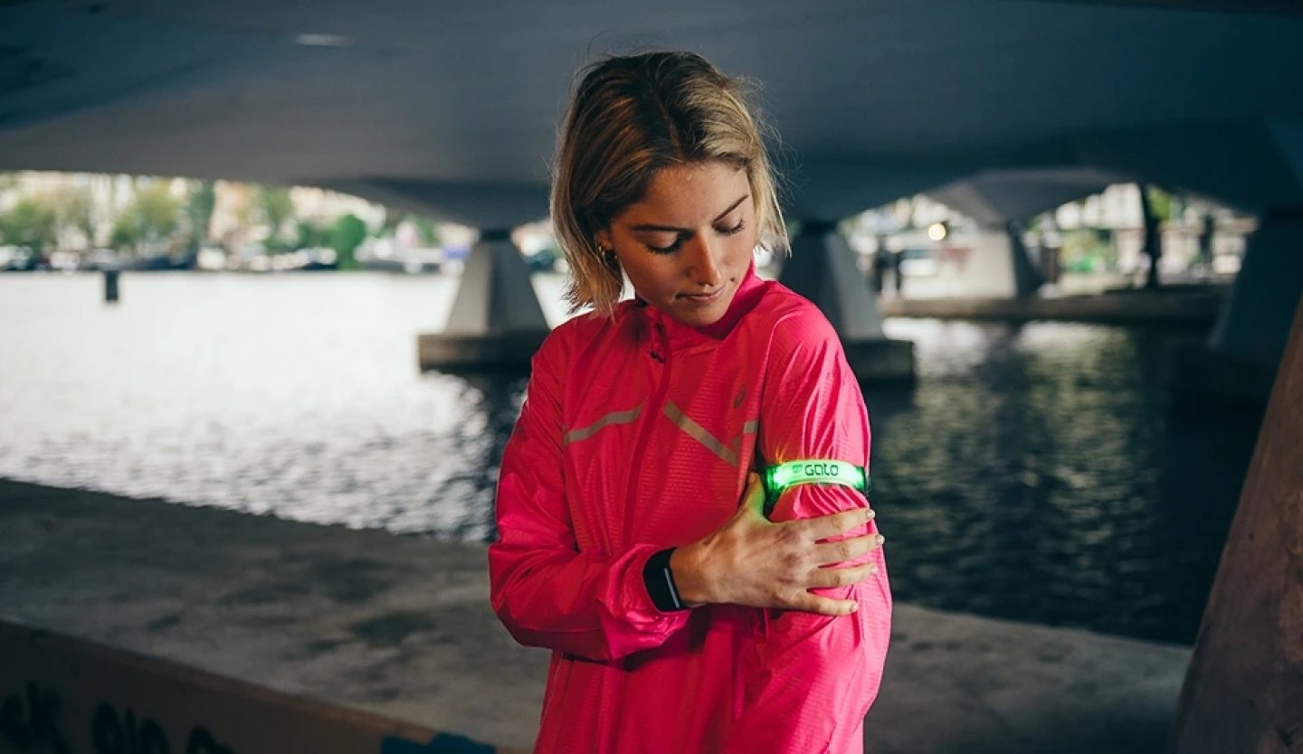 Female runner under a bridge adjusting a green LED safety arm band on her upper arm, wearing a pink reflective running jacket.