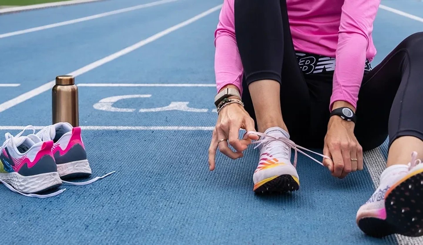 Female runner sitting on a blue athletics track tying her running spikes, with a second pair of trainers and a water bottle beside her.
