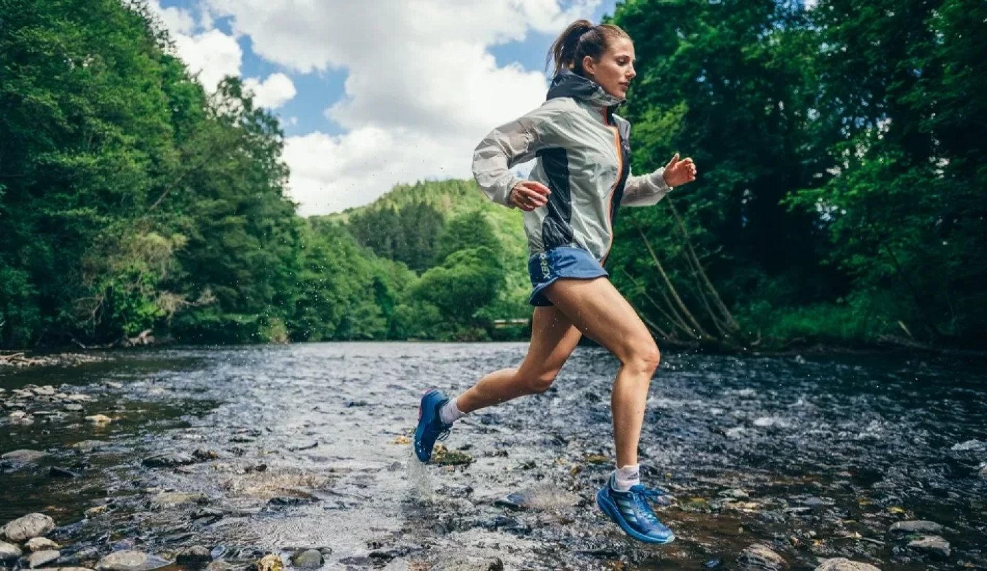 Woman trail running through a shallow rocky river wearing blue waterproof running shoes and a grey running jacket