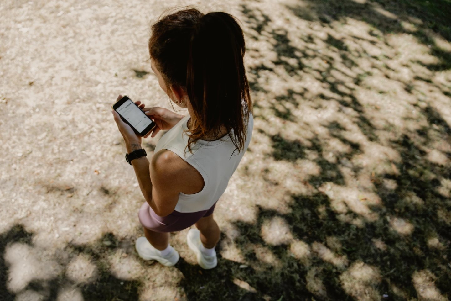 Overhead view of a female runner in white top, mauve shorts and white trainers checking her smartphone in a park.
