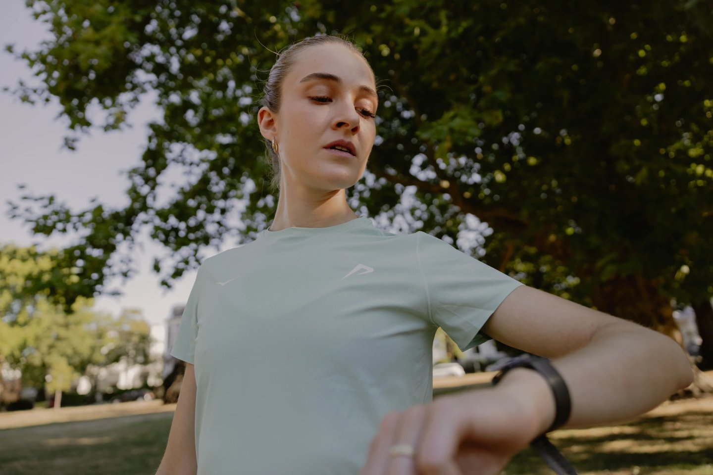 Woman in mint green short-sleeve running t-shirt checking her GPS sports watch during an outdoor park run on a sunny day.