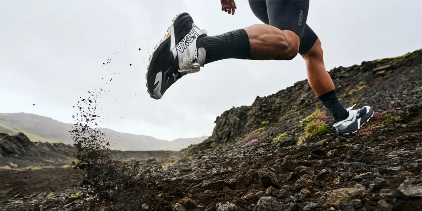 Male runner wearing HOKA trail shoes sprinting uphill over wet volcanic rock, debris flying from heel strike.