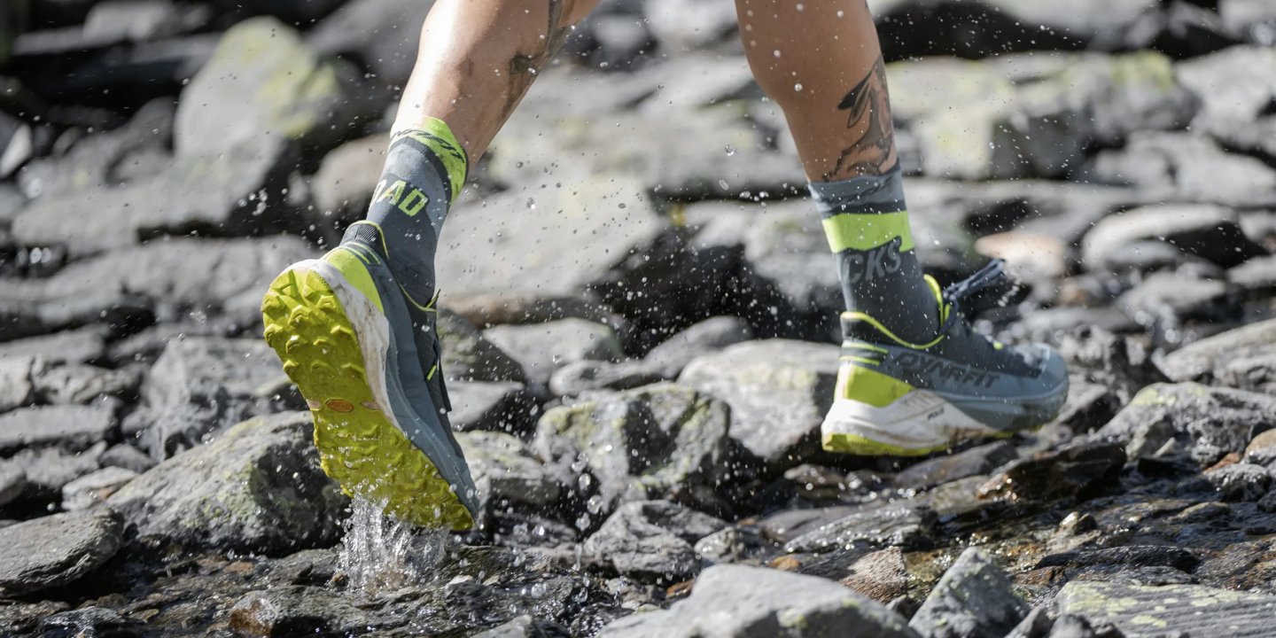 Close-up of Dynafit trail running shoes in slate blue and lime yellow splashing through a rocky stream