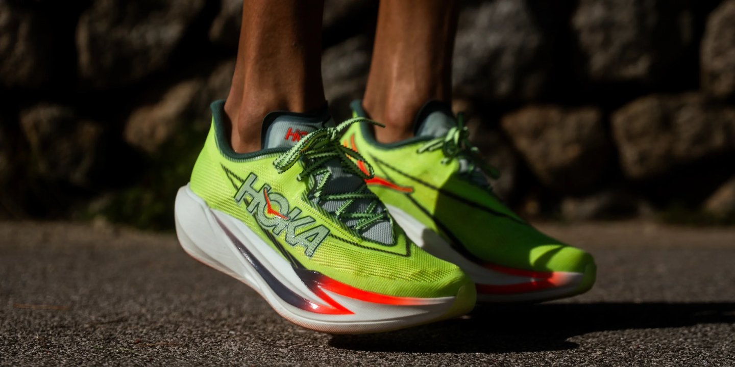 HOKA carbon-plate racing shoes in neon green with red accents and thick white rocker midsole, worn on asphalt.