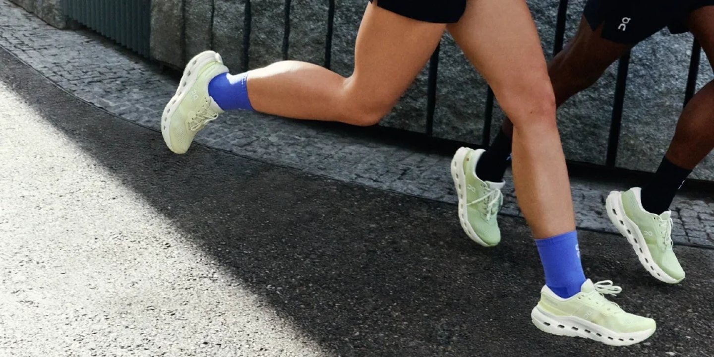 Close-up of two runners wearing lime-green On Cloudmonster shoes and blue socks sprinting on urban asphalt.