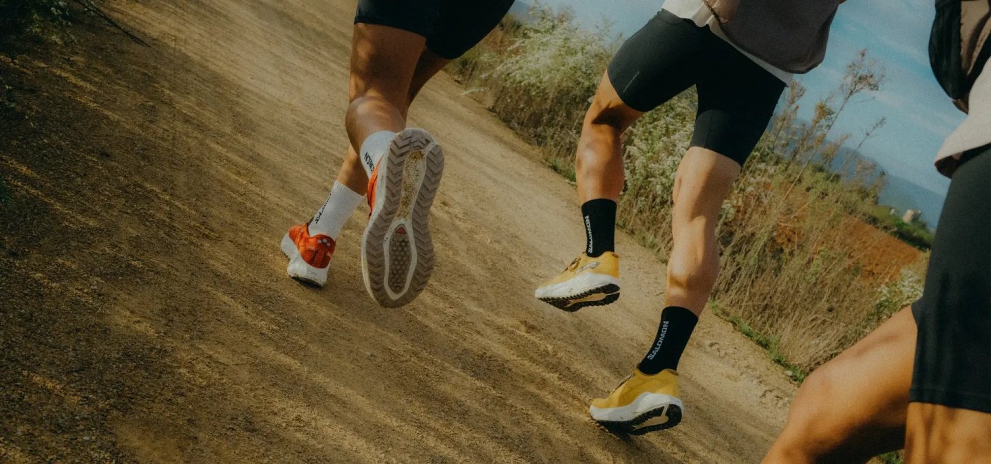 Close-up of two runners wearing Salomon trail running shoes in orange and gold on a dusty dirt path with vegetation