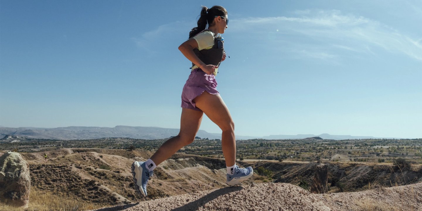 Woman trail running on rocky desert ridge wearing pink shorts, hydration vest, and Salomon trail running shoes