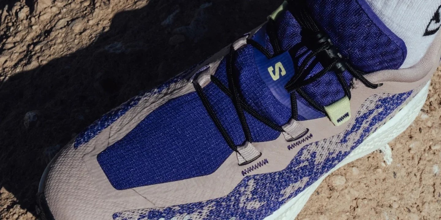 Close-up of a Salomon trail running shoe with blue knit upper, quick-lace system, and lime green accents on rocky ground.