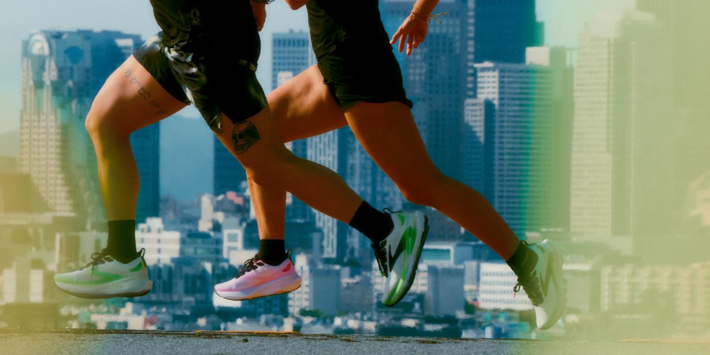 Close-up of two runners' legs mid-stride wearing green and pink performance running shoes against a city skyline backdrop