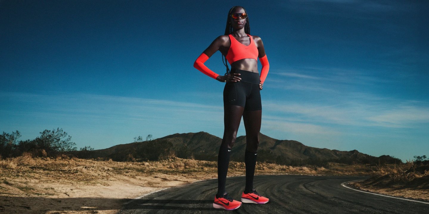 Female athlete in red Nike sports bra, black leggings, and red Nike running shoes standing on a desert road.
