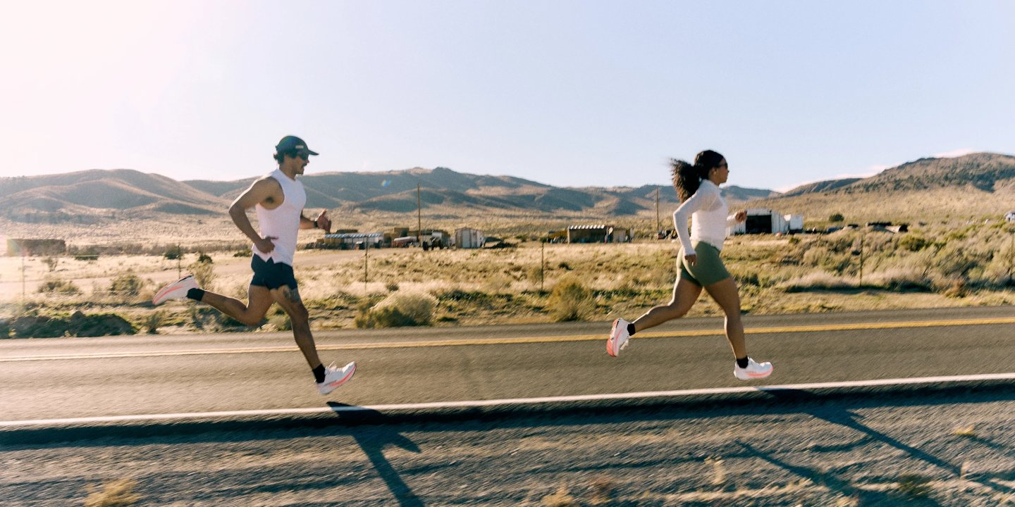 Male and female runners in white running gear and white shoes with pink soles sprinting along a desert road.