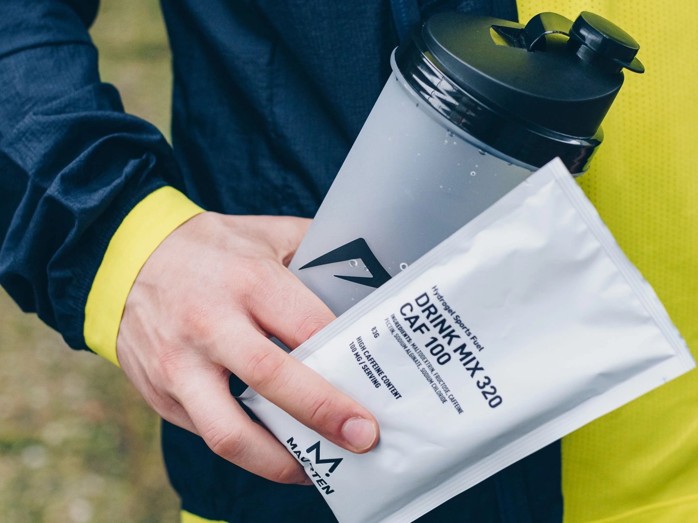 Runner holding a Maurten Drink Mix 320 CAF 100 sachet alongside a grey sports shaker bottle outdoors