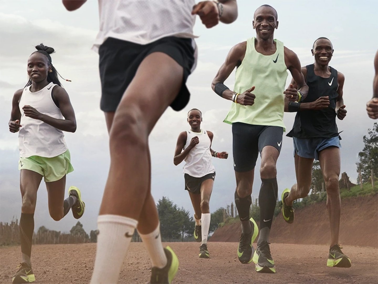 Group of elite runners wearing Nike singlets, shorts and yellow carbon-plated racing shoes running on a dirt road outdoors.
