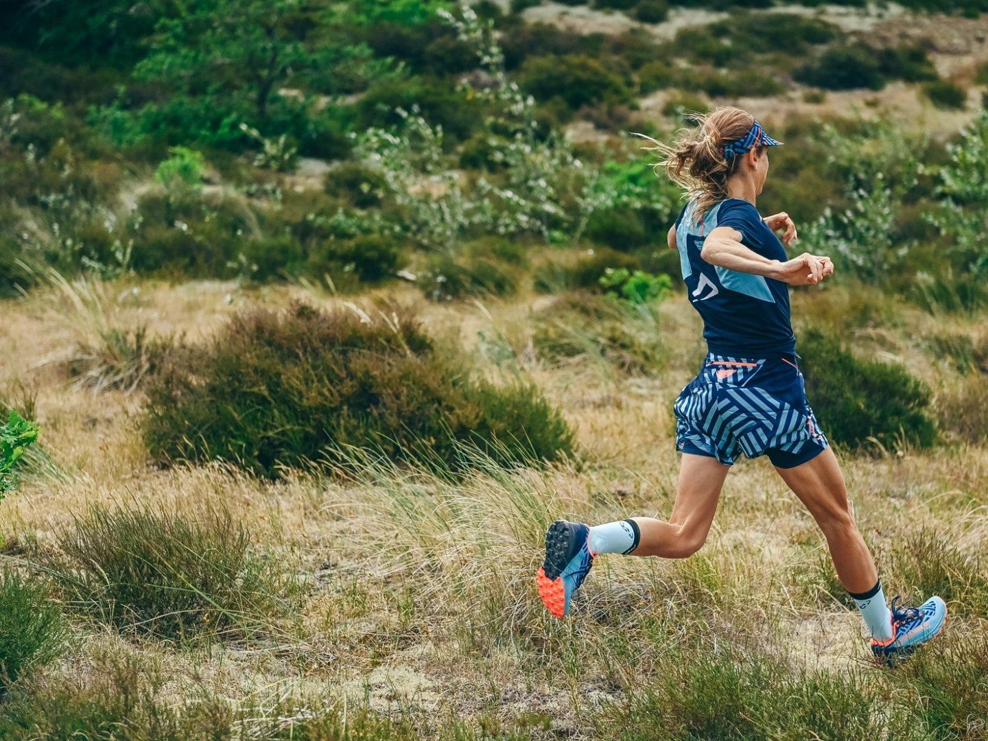 Woman trail running across open heathland wearing Carbon 2 trail shoes in blue and orange with navy patterned shorts
