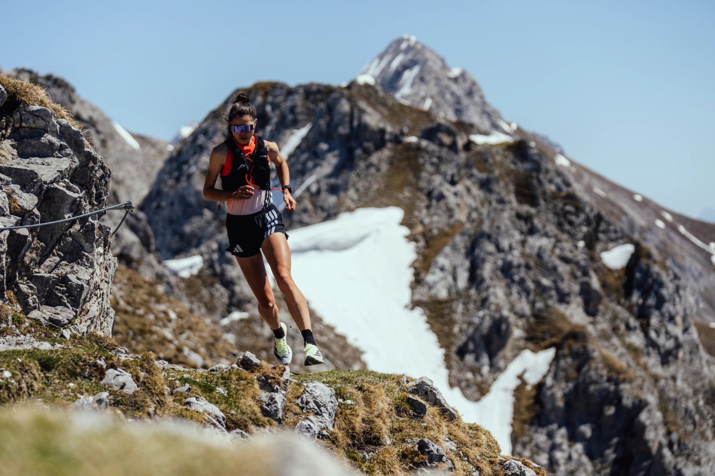 Female trail runner in black shorts, orange vest and blue sunglasses running on a rocky mountain ridge with snowy peaks behind.