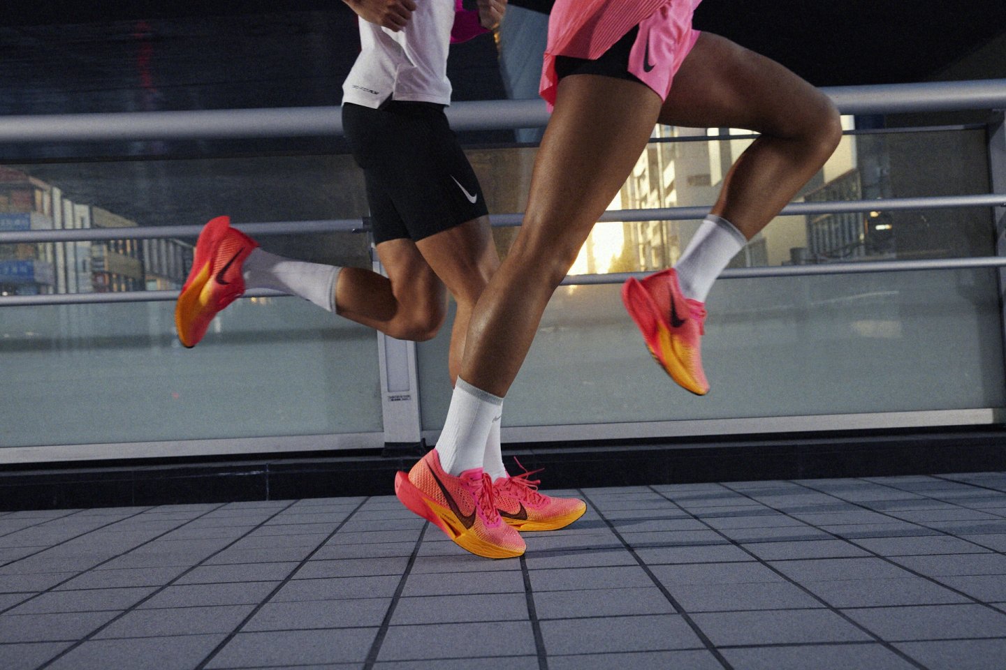 Two runners mid-stride wearing Nike ZoomX VaporFly Next% shoes in pink and orange gradient colorway on urban tiles.