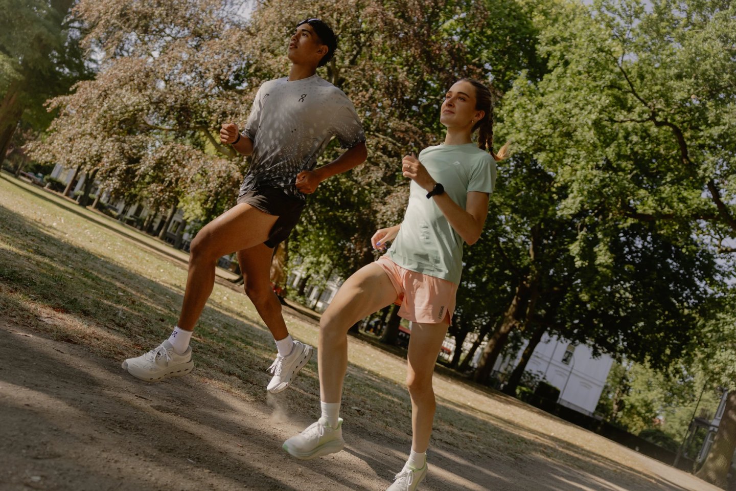 Man in grey graphic running tee and black shorts runs alongside woman in mint tee and peach shorts on a sunny park path.