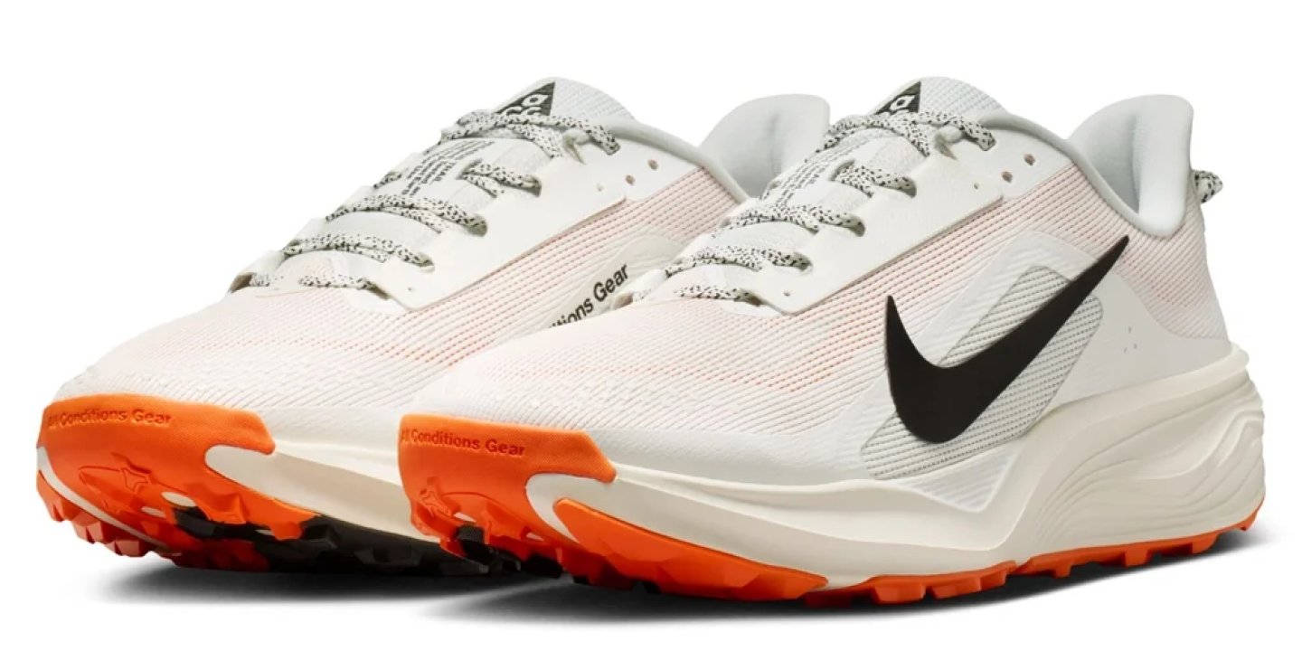 Nike ACG trail running shoes in white and cream with black Swoosh and bright orange lugged outsole.