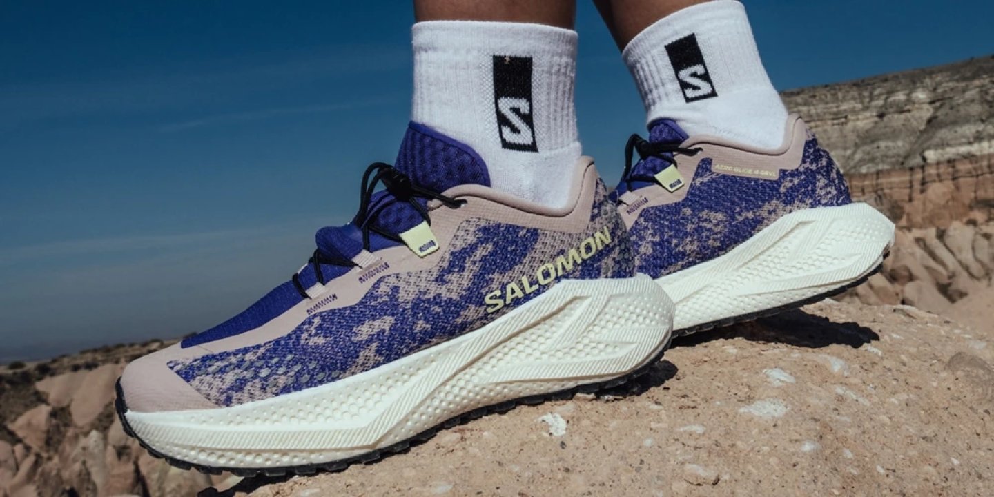 Salomon trail running shoes in blue and beige camo pattern with cream midsole worn on rocky desert cliff