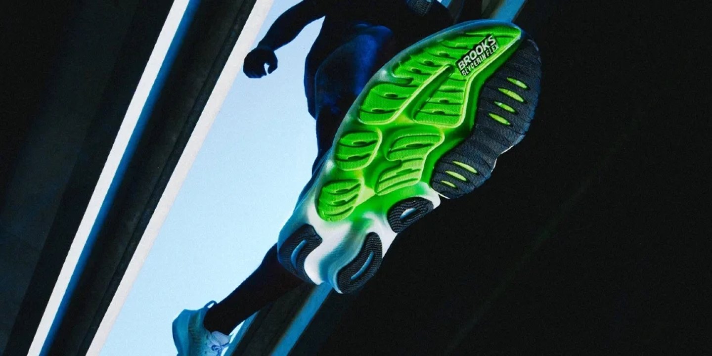Underside view of Brooks Glycerin Flex running shoe with neon green segmented outsole captured mid-stride against blue sky