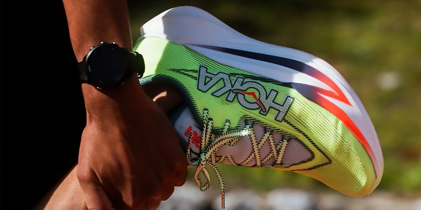 Hand holding a Hoka Rocket X2 running shoe in neon green and white with red accents and carbon plate midsole