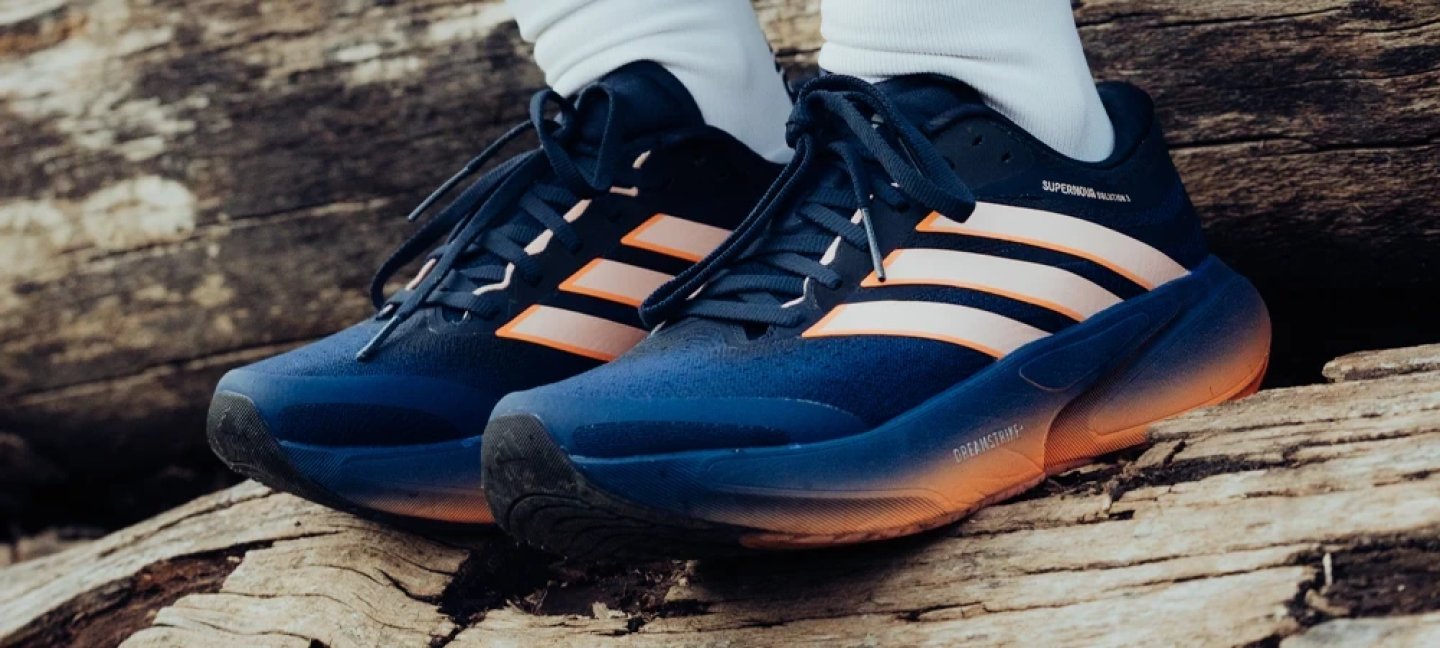 Adidas Supernova Solution 2 running shoes in navy blue and orange with white three stripes, resting on a log outdoors