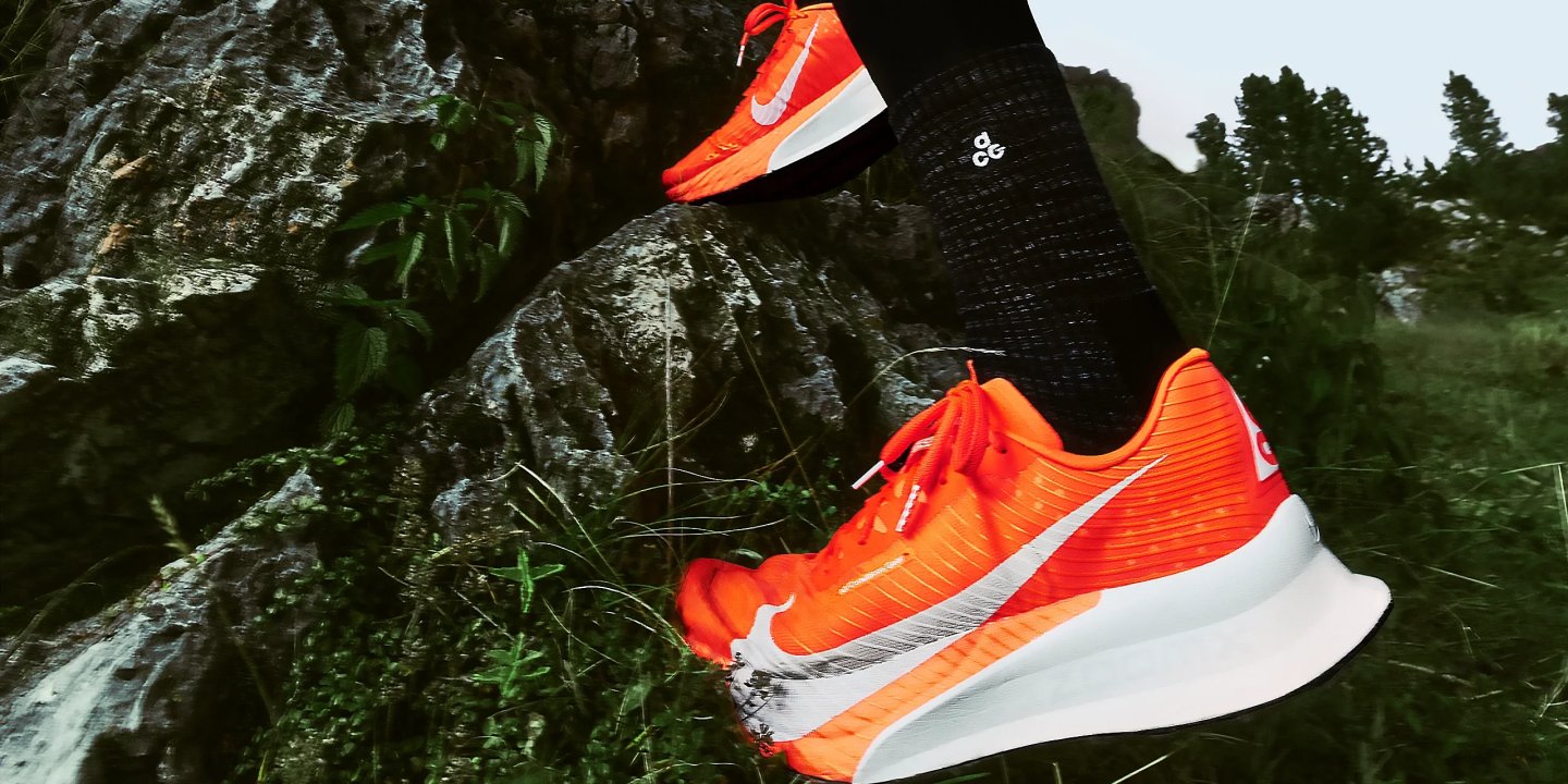 Runner wearing bright orange Nike running shoes with white Swoosh and midsole, mid-stride over rocky trail terrain.