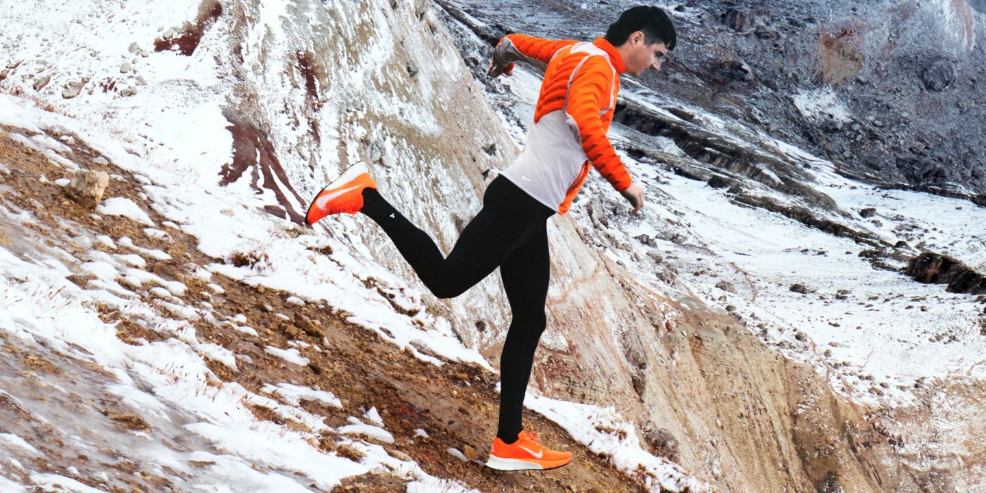 Male runner in orange Nike jacket and tights sprinting up a snow-covered rocky mountain trail.